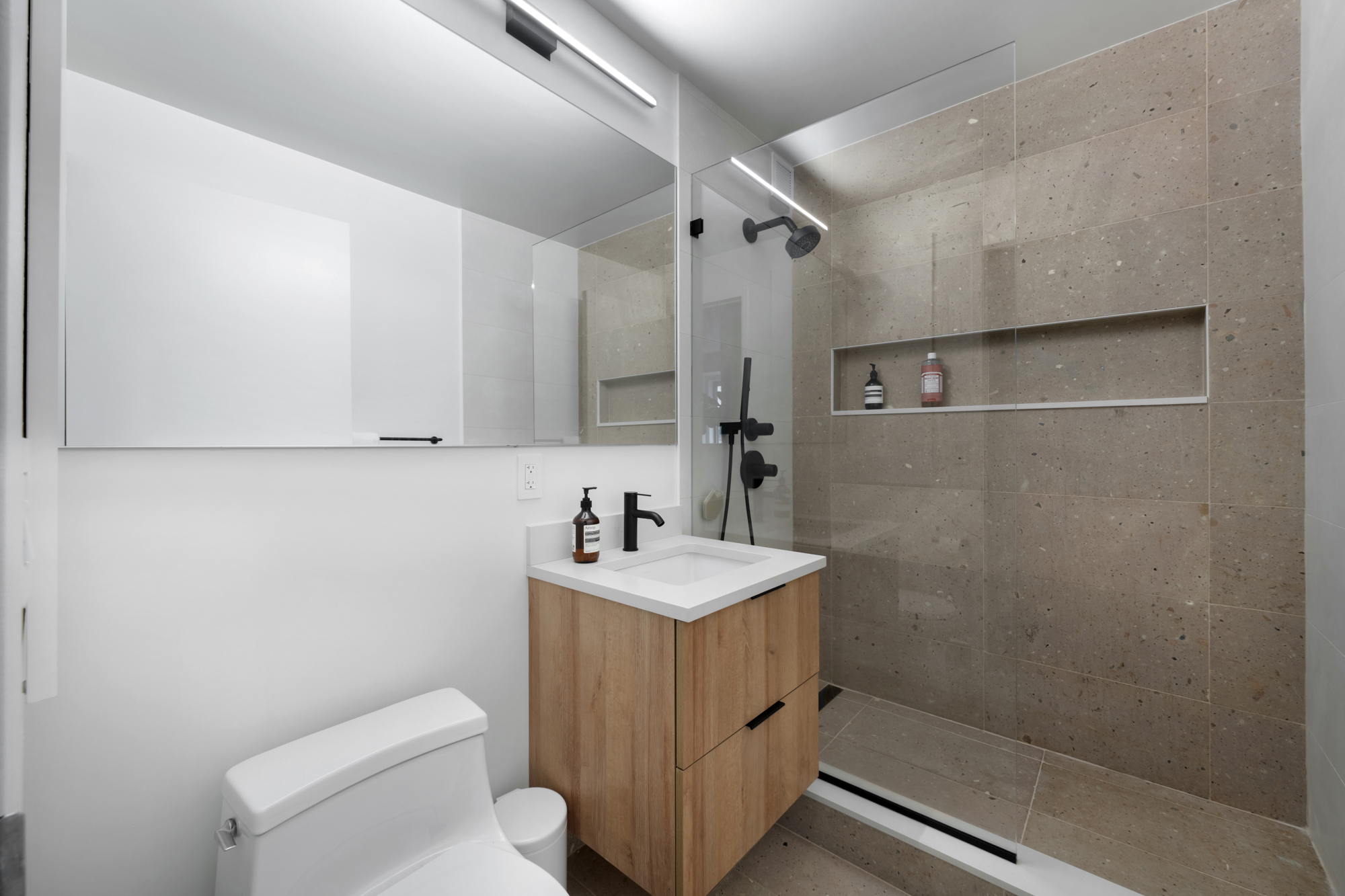 Modern NYC apartment bathroom renovation with walk-in glass shower, wood vanity, large mirror, and neutral tile