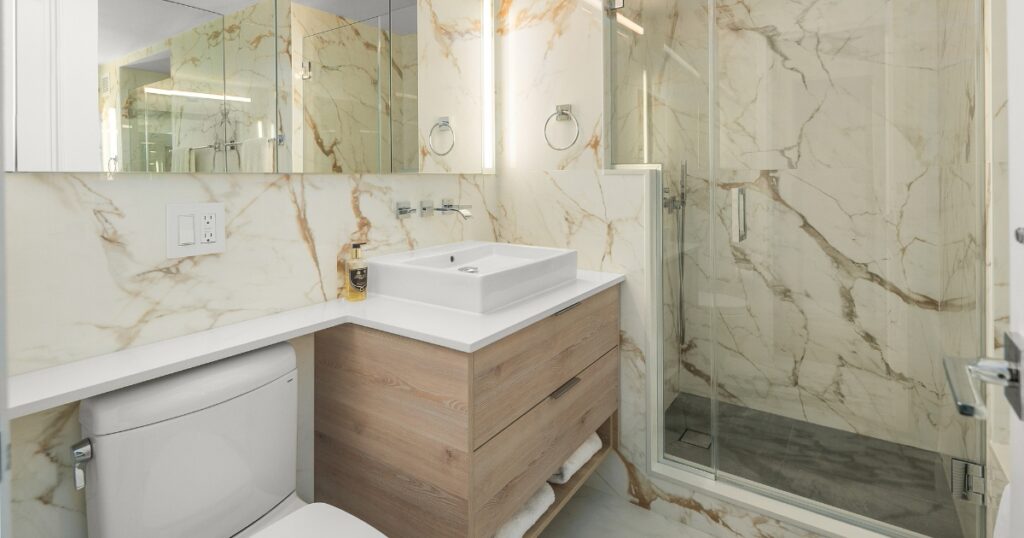 NYC apartment bathroom renovation with marble-look tile, wood vanity, glass shower, and wall-mounted faucet