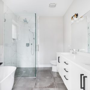 A modern, white luxury bathroom with black hardware and chrome faucets. A large glass walled shower sits in the corner with marble tiles from floor to ceiling.