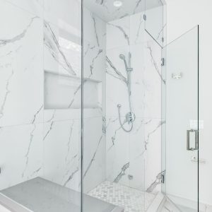 A luxurious shower with glass and marble tiled walls. The shower has a built in shelf and chrome shower head.